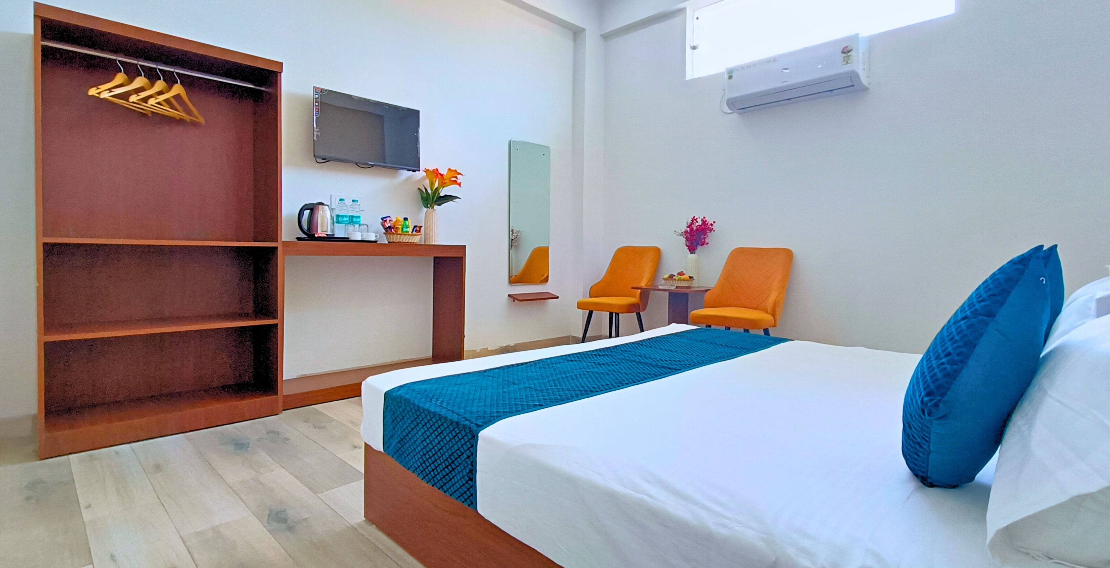 A hotel room with a bed, a desk with a chair, a TV, a wardrobe, and a small table with a vase of flowers.
