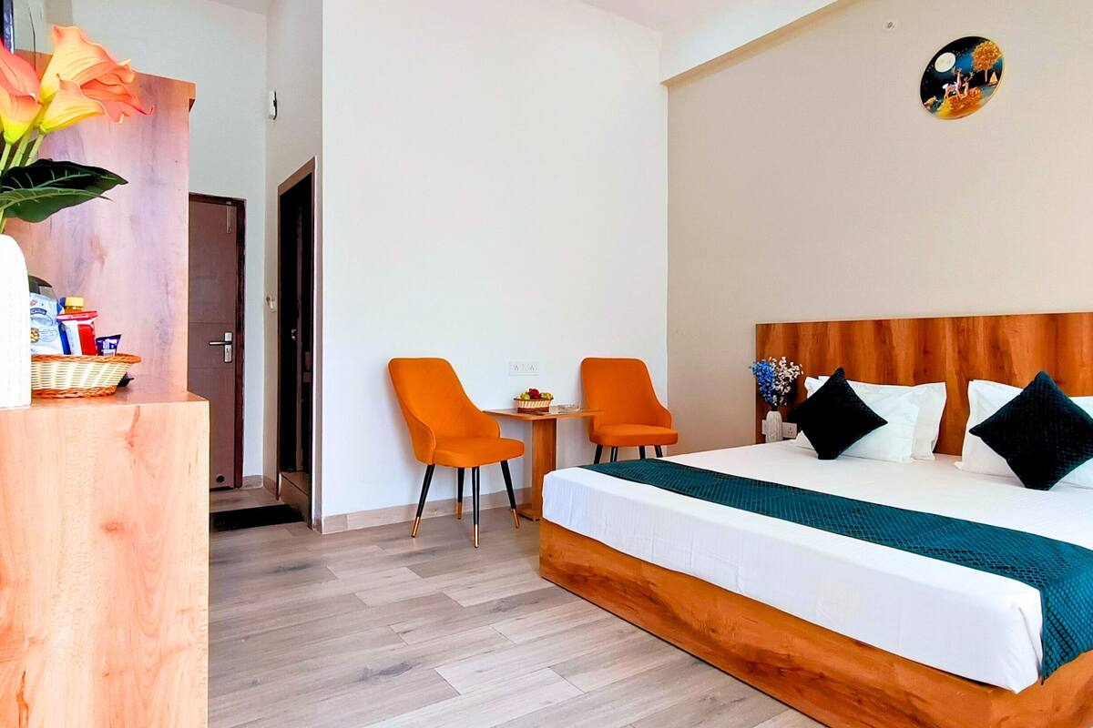 Ein Hotelzimmer mit hölzernem Kopfteil, einem großen Bett mit weißer Bettwäsche, zwei orangenen Stühlen, einem kleinen Tisch mit Obstschale und einer Vase mit Blumen auf einem Nachttisch aus Holz.
