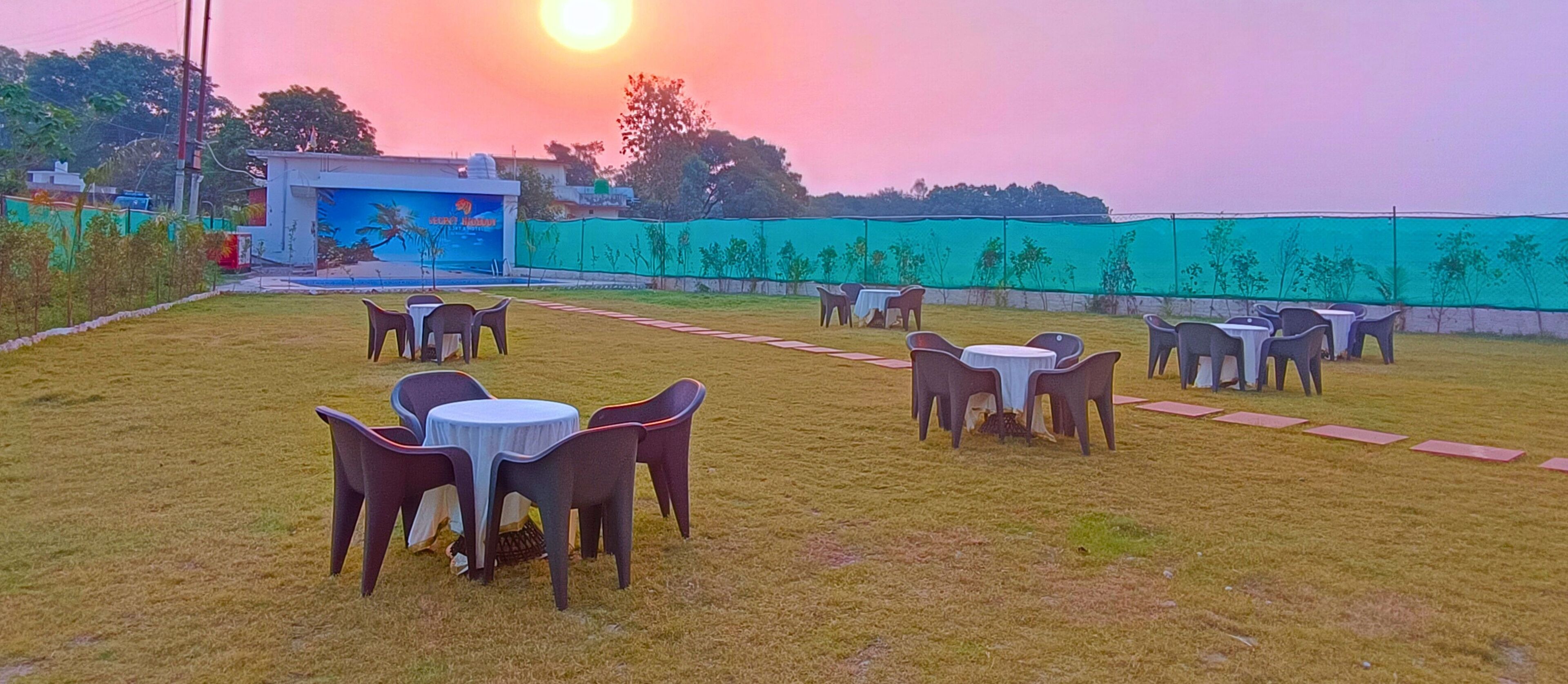 An outdoor seating area with tables and chairs, a mural on a building, and a green fence in the background.