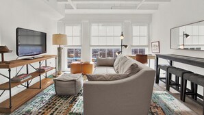 Een ruime woonkamer met een grote flatscreen-tv, een bank, voetenbankjes, een salontafel en een eethoek met barstoelen.