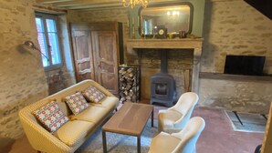A cozy room with a stone wall, a wooden sofa, a white armchair, a wooden table, and a fireplace with a wood stove.
