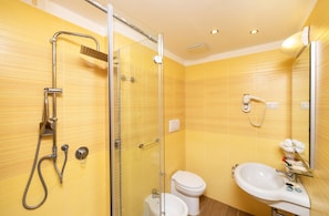 Un baño con paredes de azulejos amarillos, ducha con cerramiento de vidrio, inodoro blanco y lavamanos con artículos de aseo.