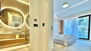 A modern hotel room with a large bed, a bathroom with a round mirror, and a view of the city through large windows. - Apartments Onia Budva (Budva)