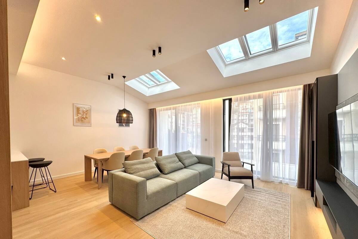 Ein modernes Wohnzimmer mit Sofa, Couchtisch und Fernseher. Es gibt Dachfenster, einen Essbereich und ein großes Fenster.