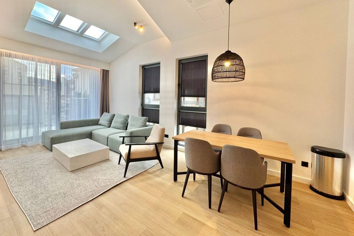 Ein modernes Wohnzimmer mit einer Couch, Sesseln, einem Esstisch und einem großen Fenster mit Jalousien.