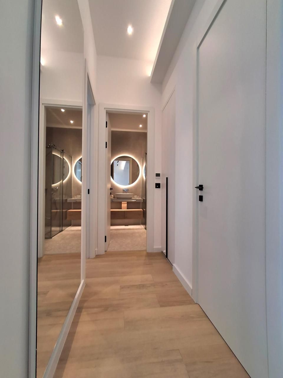 A modern hallway with wooden flooring, white walls, and a glass door leading to a bathroom with a circular mirror and a sink.