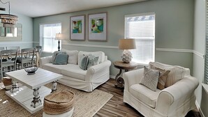 A living room with a white sofa, a coffee table, and a lamp. There are two framed pictures on the wall and a window with blinds. - Emerald Port of Call | Luxe Home, Near Beach (Miramar Beach)