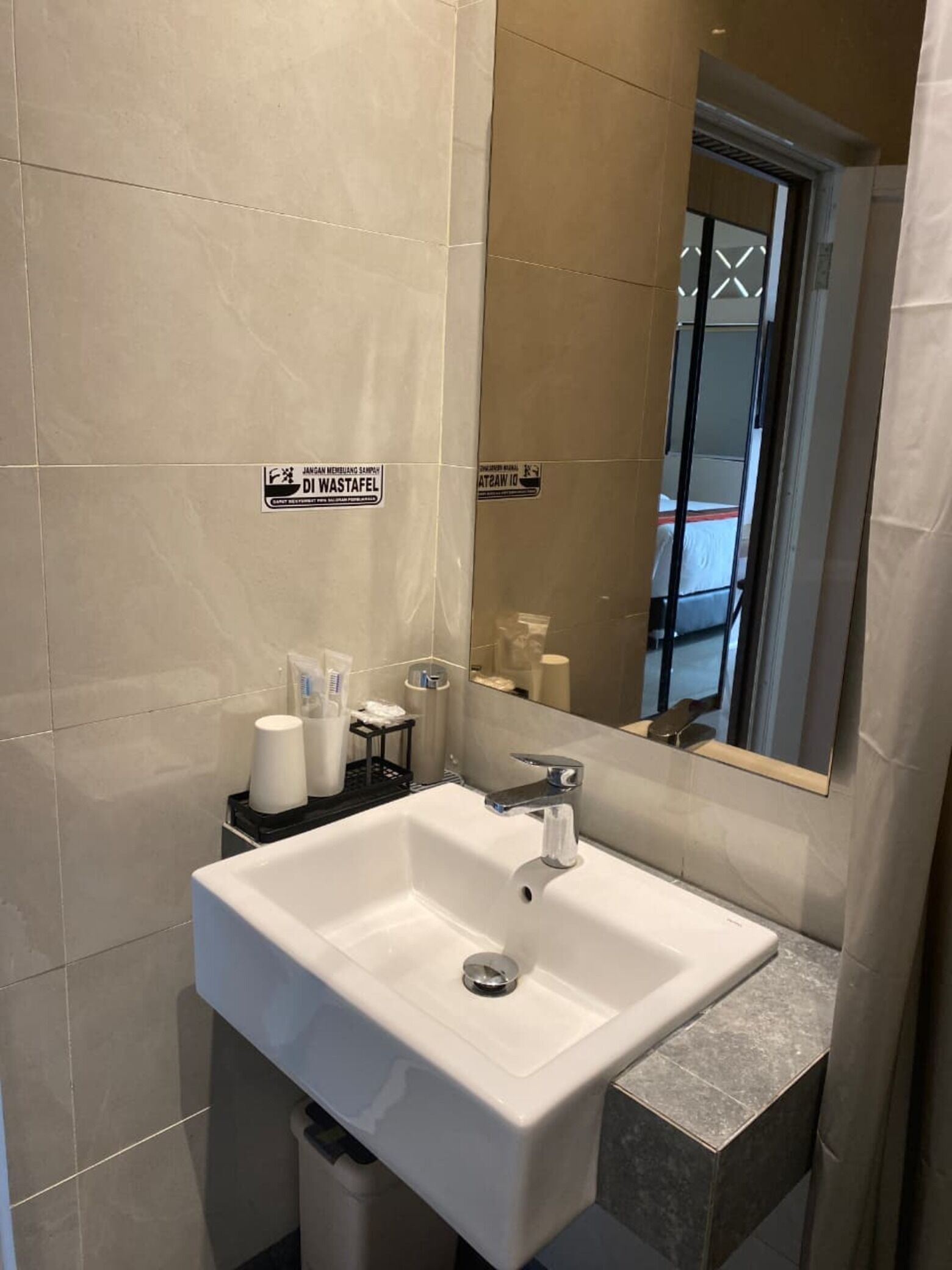 Un baño moderno con un lavamanos blanco, un espejo y un inodoro. Hay artículos de tocador sobre el lavamanos y un bote de basura debajo.