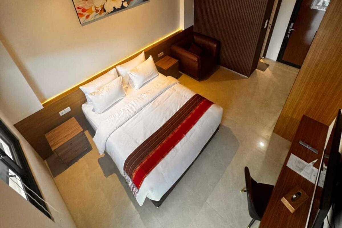 A modern hotel room with a large bed, a wooden headboard, a nightstand, a brown leather chair, a painting on the wall, and a wooden door.