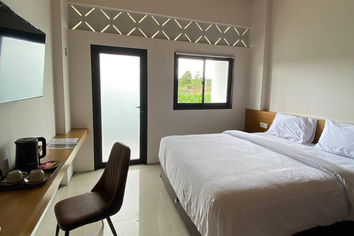 A bedroom with a bed, a chair, a desk with a coffee maker, and a window with a view of greenery.
