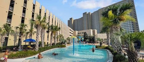 A swimming pool with a water fountain, surrounded by palm trees and lounge chairs, with tall buildings in the background.