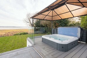 A hot tub on a wooden deck with a glass railing, overlooking a grassy area and a body of water. - Host & Stay | Bay View House (Grange-over-Sands)