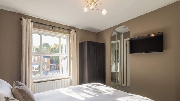 Une chambre à coucher avec un grand lit, une armoire, un téléviseur fixé au mur et une fenêtre avec des rideaux.