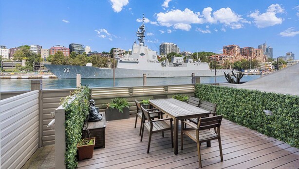 A deck with wooden furniture overlooking a body of water and a large ship.