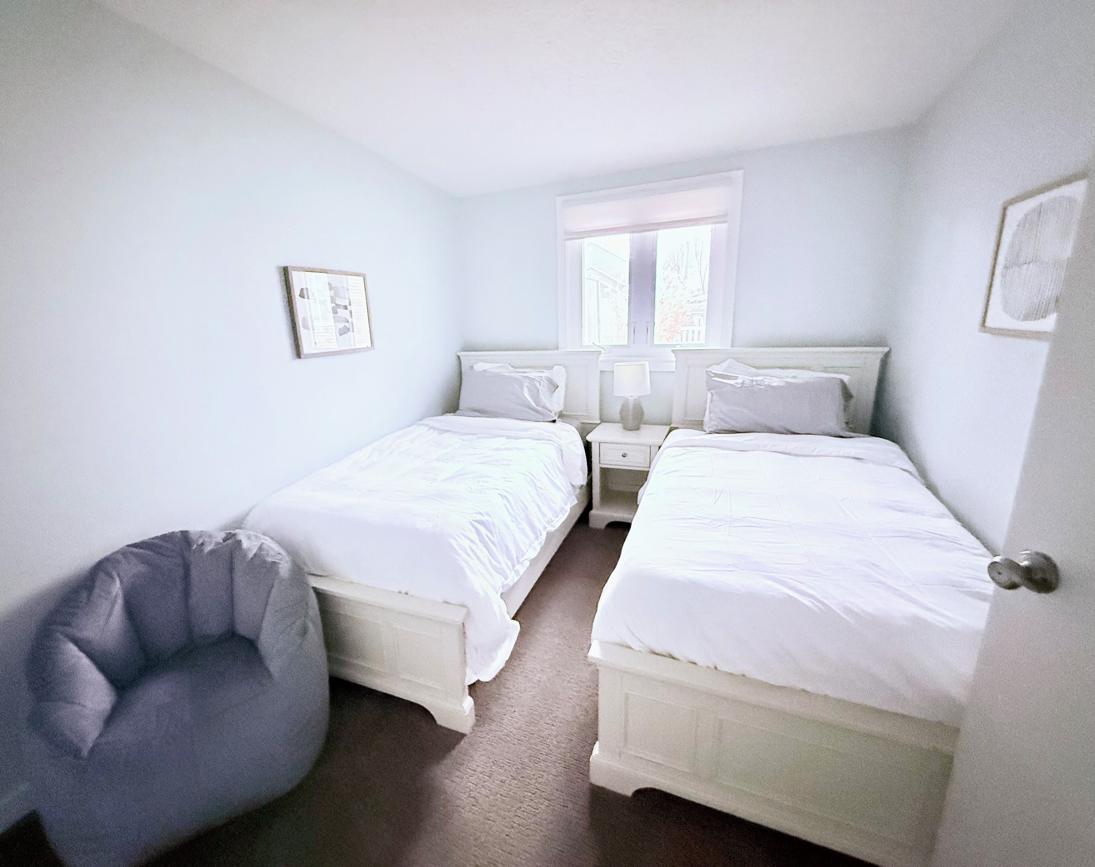 A bedroom with two single beds, a bedside table with a lamp, a chair, and a window with blinds.