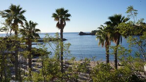 A coastal view with palm trees, a beach, and a rocky outcrop in the distance. - Enchanting beach house in Théoule-sur-Mer directly on the Plage de la Figueirette (Théoule-sur-Mer)
