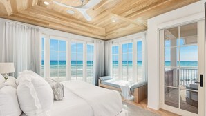 Un dormitorio con una cama grande, ventilador de techo y vistas a la playa a través de amplias ventanas.