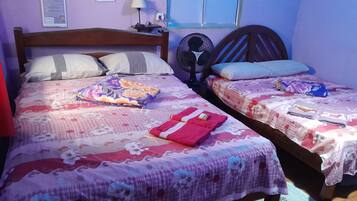 A room with two beds, each with floral bedding and pillows. There is a fan on one bed and a lamp on the other.