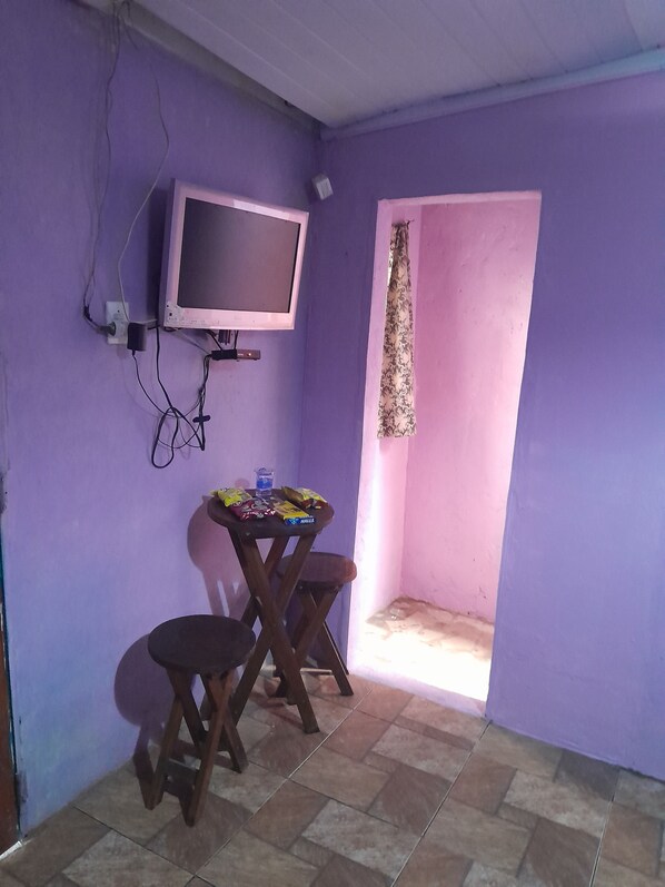 A small room with purple walls, a tiled floor, a television mounted on the wall, a small table with chairs, and a door leading outside. - Hospedagem Chapéu Virado  (Belém)