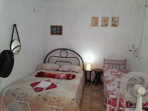 A bedroom with a bed, a bedside table with a lamp, a fan, and a small shelf with a flower. - Hospedagem Chapéu Virado  (Belém)