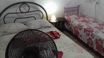 A bedroom with a bed, a standing fan, a bedside table with a lamp, and a small shelf with a flower.