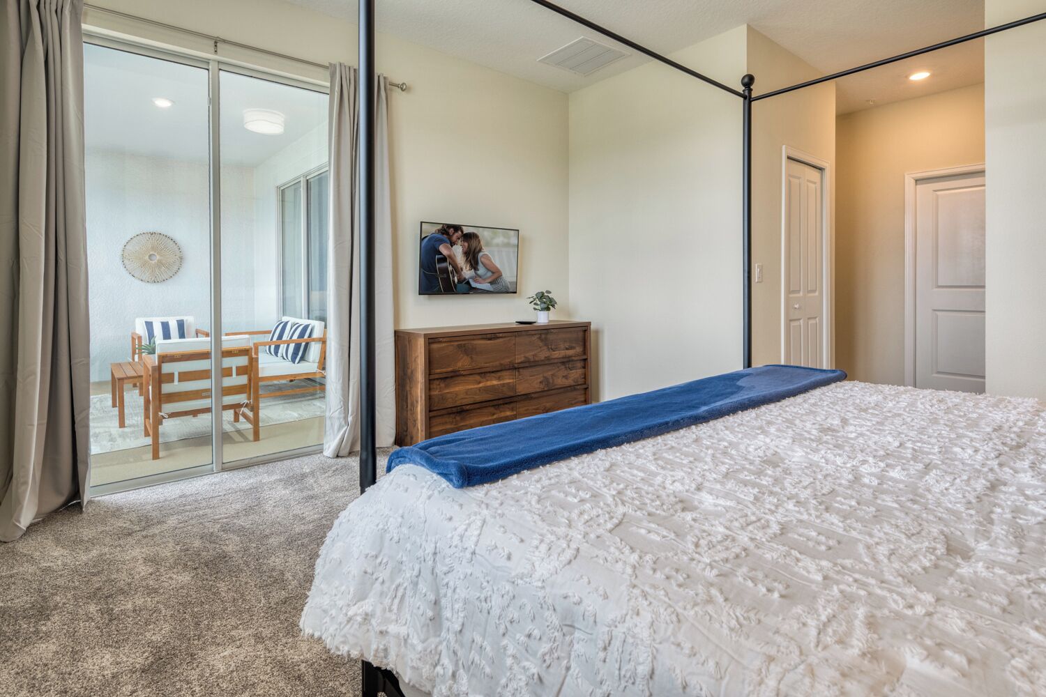 Une chambre avec un grand lit, une commode en bois, un téléviseur fixé au mur et une porte-fenêtre coulissante donnant sur un balcon équipé d’une chaise et d’une table.