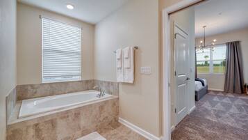 A modern bathroom with a large bathtub, a window with blinds, and a door leading to a bedroom.