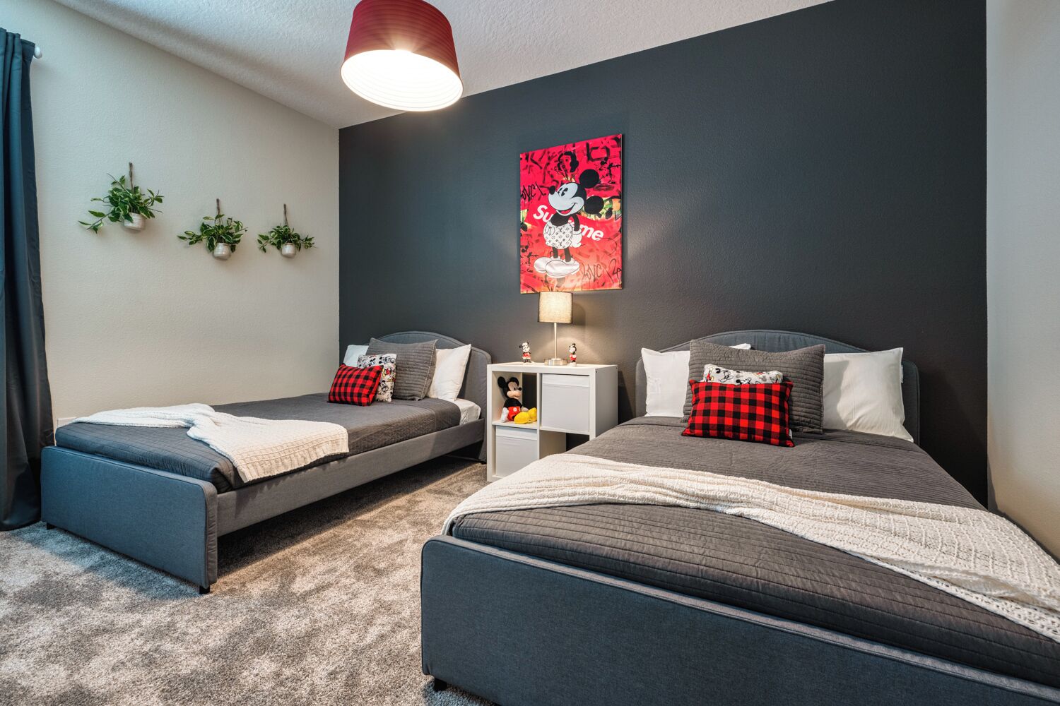 Une chambre avec deux lits, une table de chevet, une affiche de Mickey Mouse et trois plantes en pot.