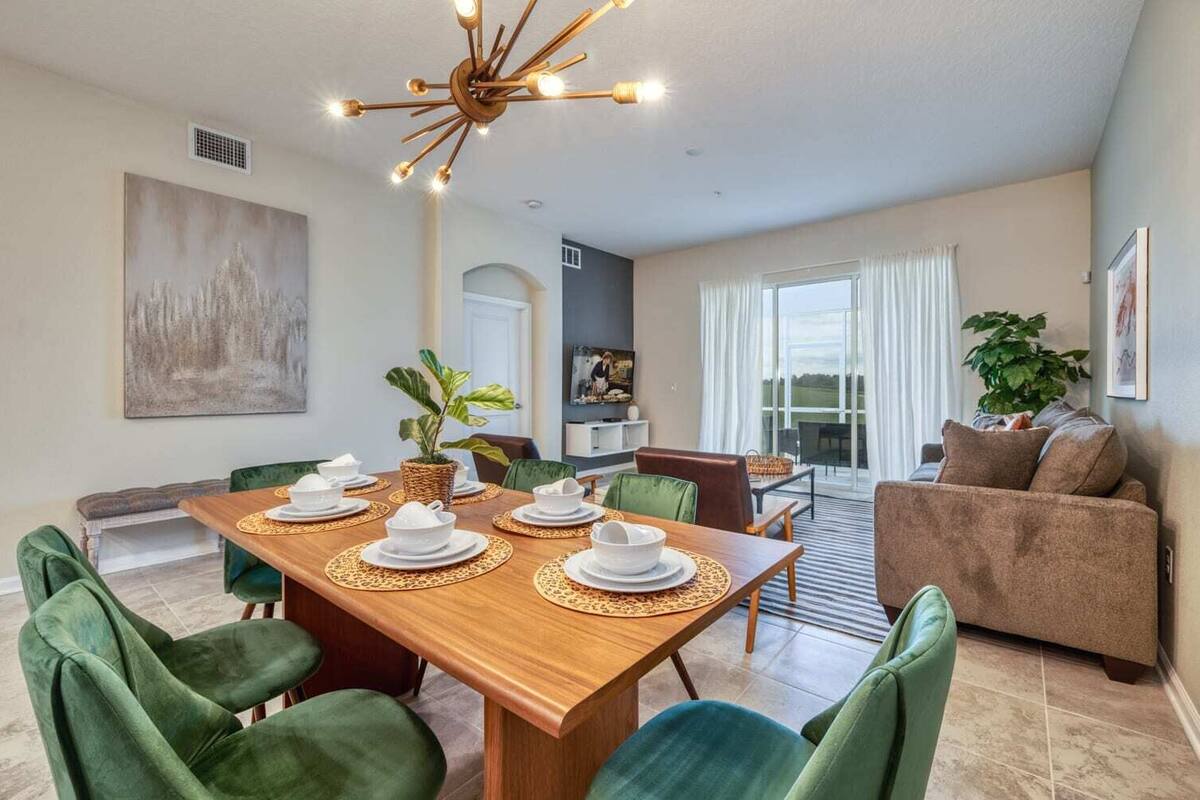 Un coin repas avec une table en bois dressée pour quatre personnes, des chaises à l’assise rembourrée de tissu vert et un lustre moderne.