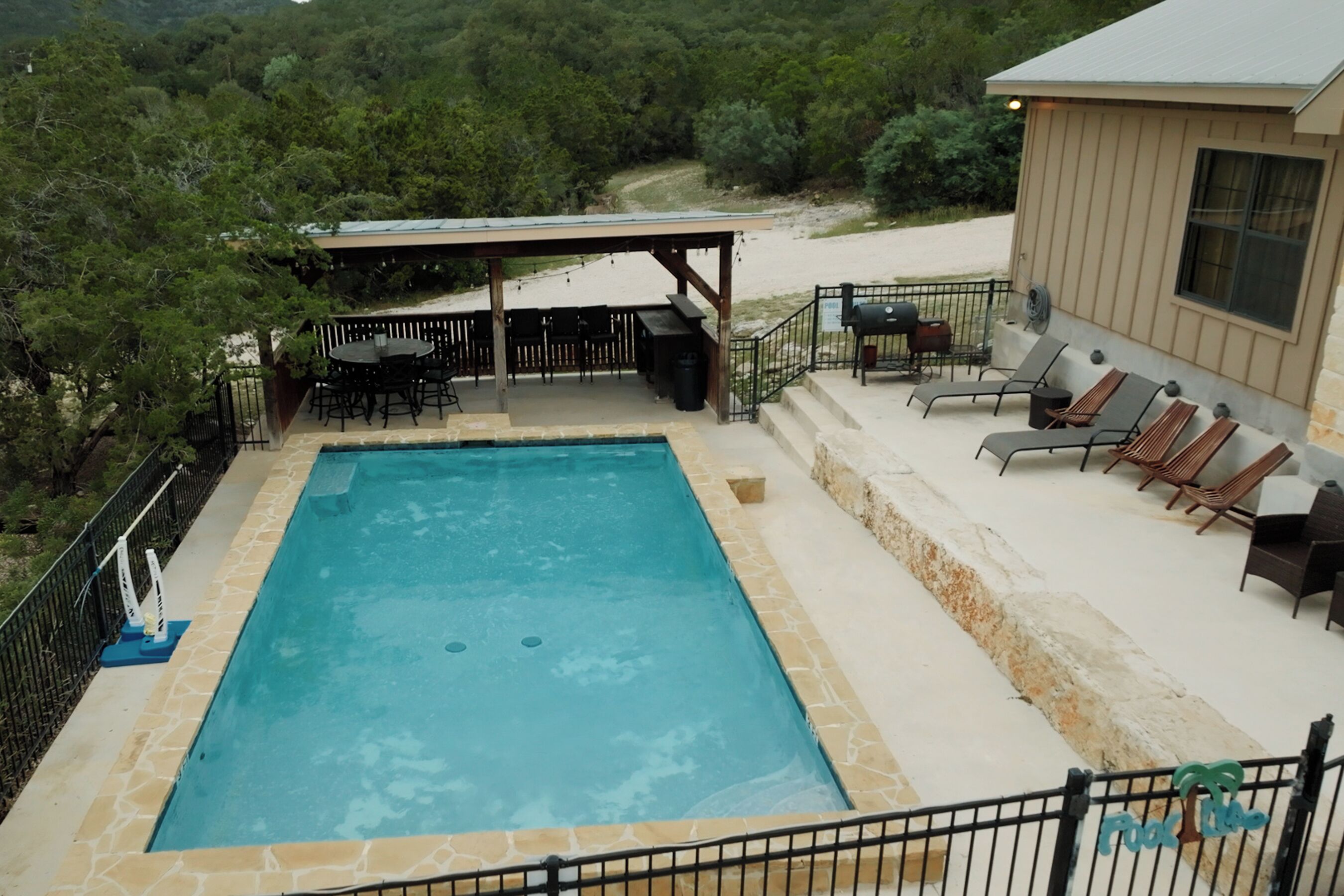 A rectangular pool with a stone border, surrounded by a patio area with outdoor furniture and a covered seating area.