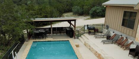 A rectangular pool with a stone border, surrounded by a patio area with outdoor furniture and a covered seating area.