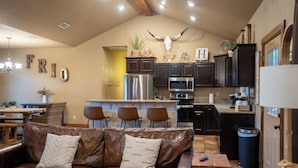 A spacious living room with a leather sofa and a dining area with a wooden table and chairs. A kitchen with dark cabinets and stainless steel appliances is visible in the background.