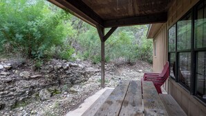 A covered outdoor seating area with a wooden bench and a red chair, overlooking a rocky stream and dense greenery.