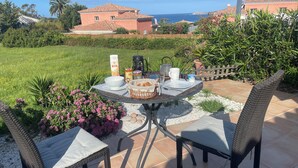 Un patio con una mesa redonda dispuesta para el desayuno, rodeada de sillas de mimbre, y una vista panorámica de casas y el mar al fondo.