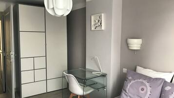 Une chambre moderne avec un bureau en verre, une chaise blanche, un lit avec un oreiller violet, une lampe fixée au mur et un tableau encadré.