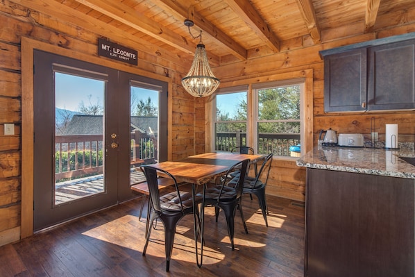 A wooden cabin interior with a dining table, chairs, a chandelier, and a kitchen area.
