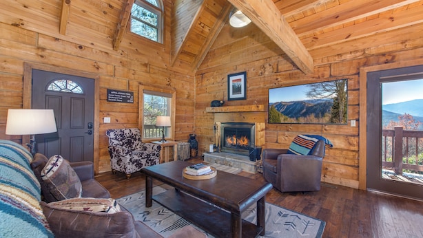 Ein rustikales Blockhaus-Interieur mit Kamin, Holzwänden, einer Couch, einem Couchtisch und Bergblick durch ein großes Fenster.
