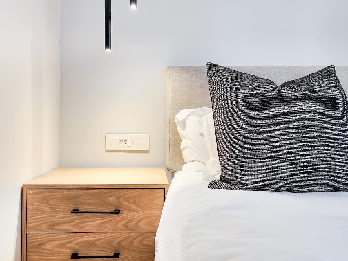 A neatly made bed with a white comforter and a patterned pillow, a wooden nightstand with a drawer, and a wall socket.