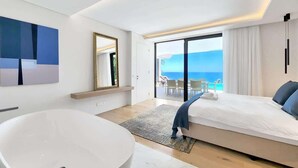 Une chambre d’hôtel moderne dotée d’une grande baignoire, d’un lit avec vue sur la mer et d’un balcon avec coin salon extérieur.
