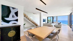 Un espace repas moderne avec une table et des chaises en bois, un escalier avec des marches en bois et une grande fenêtre offrant une vue sur l’océan.
