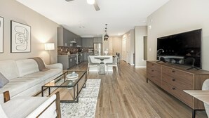 A modern living room with a sofa, coffee table, and TV. There is a dining area with a table and chairs, and a kitchen in the background. - Landing | Modern 1BD, Clubhouse, Yoga Studio (Phoenix)