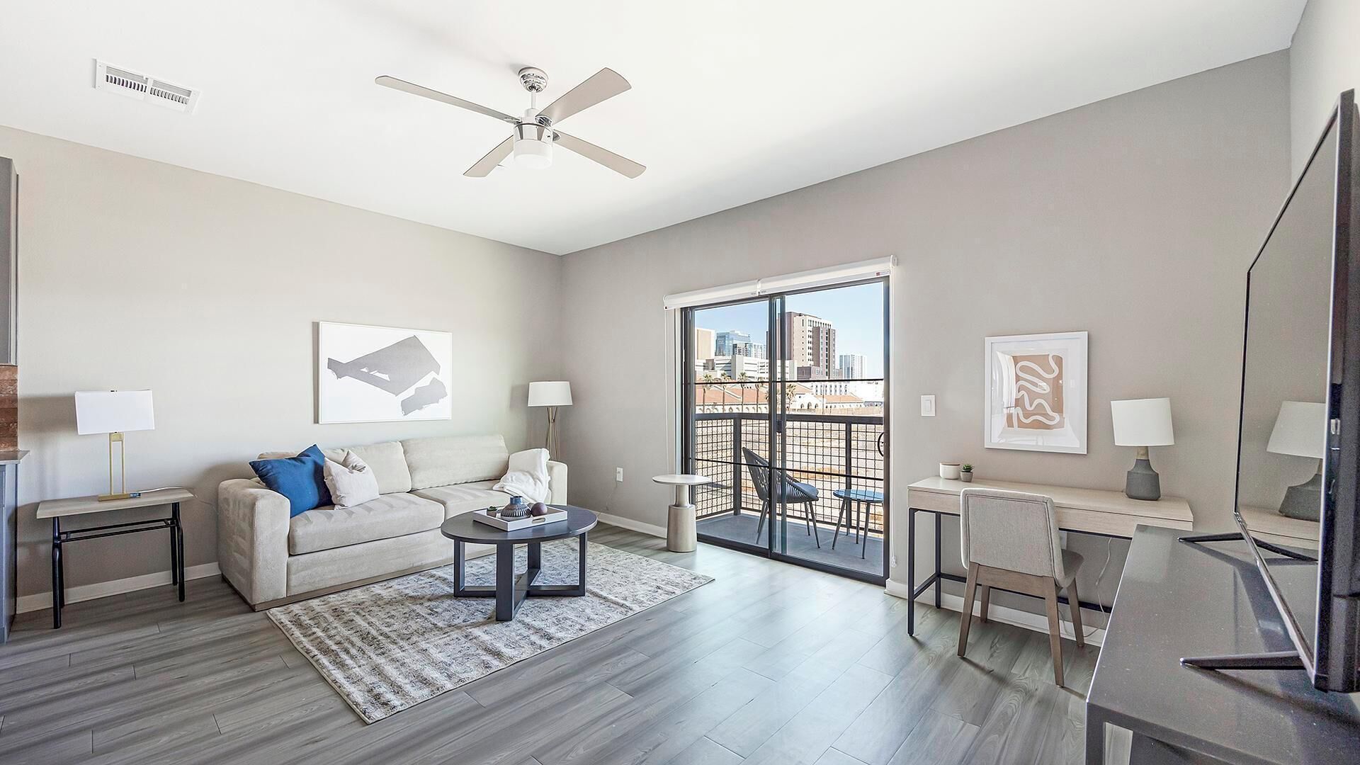 Ein modernes Wohnzimmer mit Sofa, Couchtisch und Schreibtisch mit Stuhl. Es gibt einen Deckenventilator, ein Fenster mit Blick auf Gebäude und eine Tür, die auf einen Balkon führt.