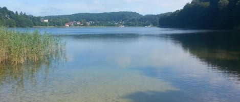 A calm lake with clear water, surrounded by forested hills and a few buildings in the distance.