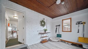 Una habitación con paredes de ladrillo blanco, techo de madera, una cama para mascotas, una pequeña mesa con una planta y utensilios de limpieza en la pared.
