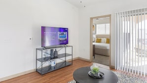 A modern hotel room with a flat-screen TV on a glass shelf, a round table with a plant, and a sliding glass door leading to a balcony. - Landing | Stylish 1BD, Rooftop Lounge, Gym (Fort Worth)
