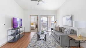 A modern living room with a flat-screen TV, a coffee table, a sofa, and a view through a glass door to an outdoor balcony. - Landing | Stylish 1BD, Rooftop Lounge, Gym (Fort Worth)
