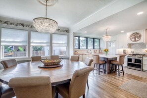 A spacious dining area with a wooden table and chairs, a chandelier, and a view of the backyard through large windows. - DOWNTOWN BRADENTON RETREAT | SWIM SPA • AMI • IMG (Bradenton)