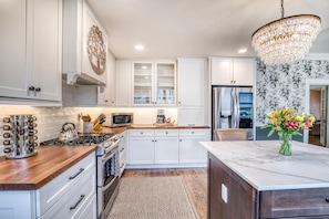 A modern kitchen with white cabinetry, a central island, a stainless steel refrigerator, and a chandelier. - DOWNTOWN BRADENTON RETREAT | SWIM SPA • AMI • IMG (Bradenton)