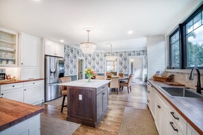 A modern kitchen with white cabinetry, a central island, and a dining area with a chandelier. - DOWNTOWN BRADENTON RETREAT | SWIM SPA • AMI • IMG (Bradenton)