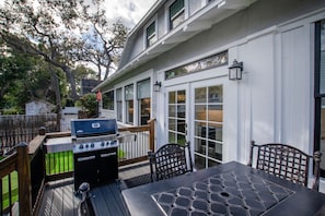 A deck with a grill, outdoor table, and chairs, overlooking a backyard with a fence and trees. - DOWNTOWN BRADENTON RETREAT | SWIM SPA • AMI • IMG (Bradenton)